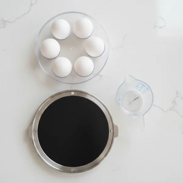 Disassembled egg cooker showing heating plate, egg rack, measuring cup with piercing pin.